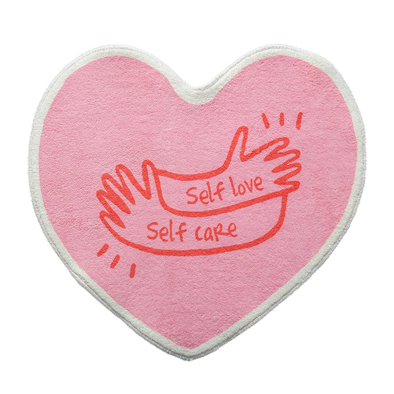 Tappeto da camera rosa a forma di cuore per ragazze. Sulla parte superiore, una stampa di due mani con le parole self love self care in rosso.
