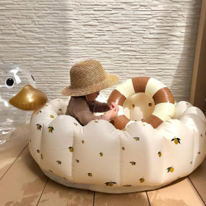 Piscina per bambini gonfiabile bianca con motivi di ulivi, posta su un pavimento di legno di fronte a una parete bianca con all'interno un bambino con un cappello di paglia