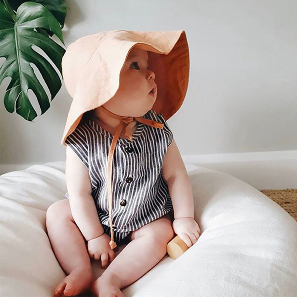Bambino seduto su un cuscino bianco con un cappello da sole beige a tesa larga e un vestitino a righe davanti a una parete grigio chiaro con una foglia di pianta verde.