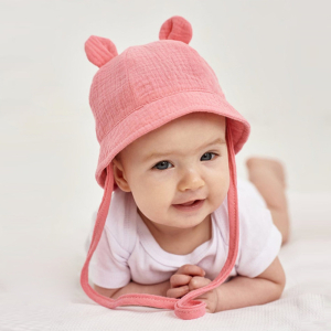 Bambina sdraiata su un tessuto bianco a nido d'ape con un body bianco e un cappello di mussola rosa con orecchie e cordoncino.