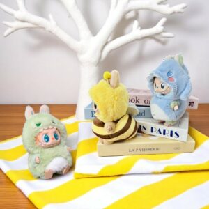 3 peluche Labubu con costumi di animali in peluche, disposte su un asciugamano giallo e bianco, su un tavolo di legno con un albero bianco sullo sfondo