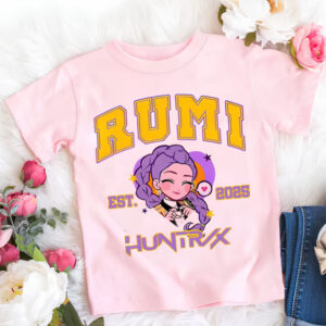 T-shirt per bambina K-pop Demon Hunters rosa del personaggio Rumi con il suo nome scritto in maiuscolo giallo così come Huntrix. Tra i due, il viso versione cartoon di Rumi. La t-shirt è posata accanto ai fiori.